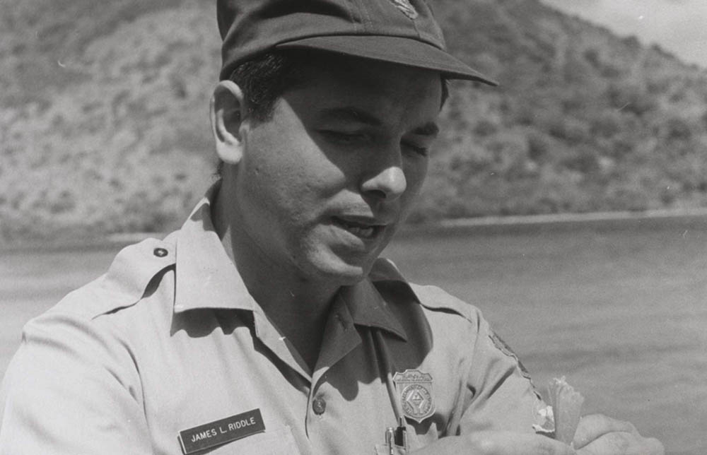 James Riddle in NPS uniform with ball cap wears a shield-shaped badge featuring stylized hands.